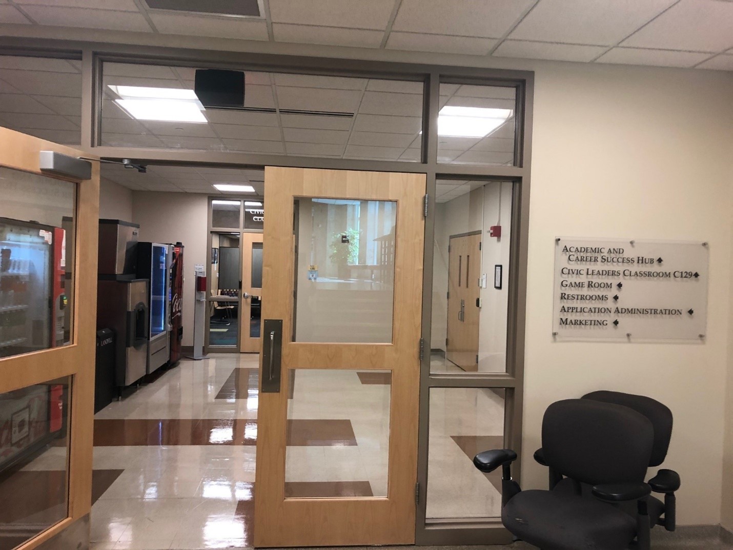 A set of clear double doors inside the Briscoe Quadrangle. There is a sign with directions on the wall next to the doors, and past the doors is a row of vending machines.